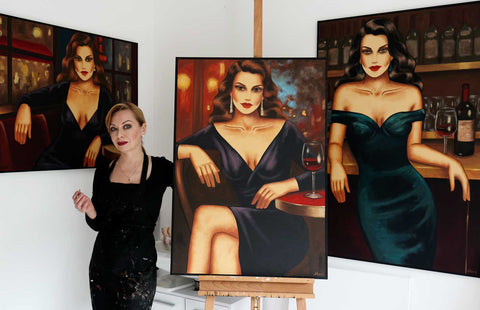 Künstlerin Ekaterina Moré mit drei Weinbildern eleganter Frauen an einer Bar in ihrem Atelier